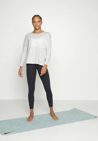 Donna in piedi a piedi nudi su un tappetino per esercizi blu, indossa una maglietta bianca a maniche lunghe e leggings neri, su uno sfondo bianco uniforme.