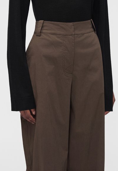 Gros plan d'une personne portant un pantalon marron taille haute et ample ainsi qu'un haut noir à manches longues, les mains détendues sur les côtés.