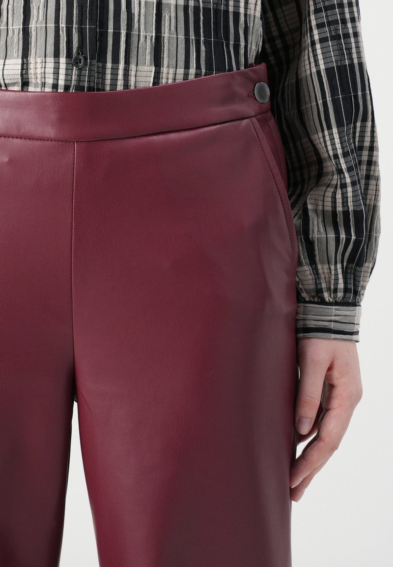 Bordeauxfarbene Kunstledershorts mit glatter Textur, hoher Taille und einem schwarzen Knopfverschluss. Kombiniert mit einer schwarz-weißen gestreiften Bluse.