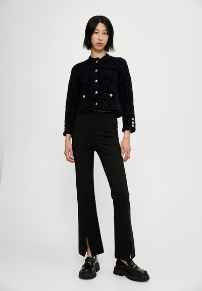 Chaqueta de textura navy con cierres de botones dorados, combinada con pantalones negros ajustados con aberturas en el dobladillo y zapatos negros gruesos.