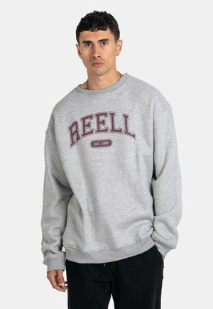 Grau Sweatshirt aus weichem Stoff, mit großem burgunderrotem "REELL"-Schriftzug und Rundhalsausschnitt. Dazu schwarze Cordhosen.