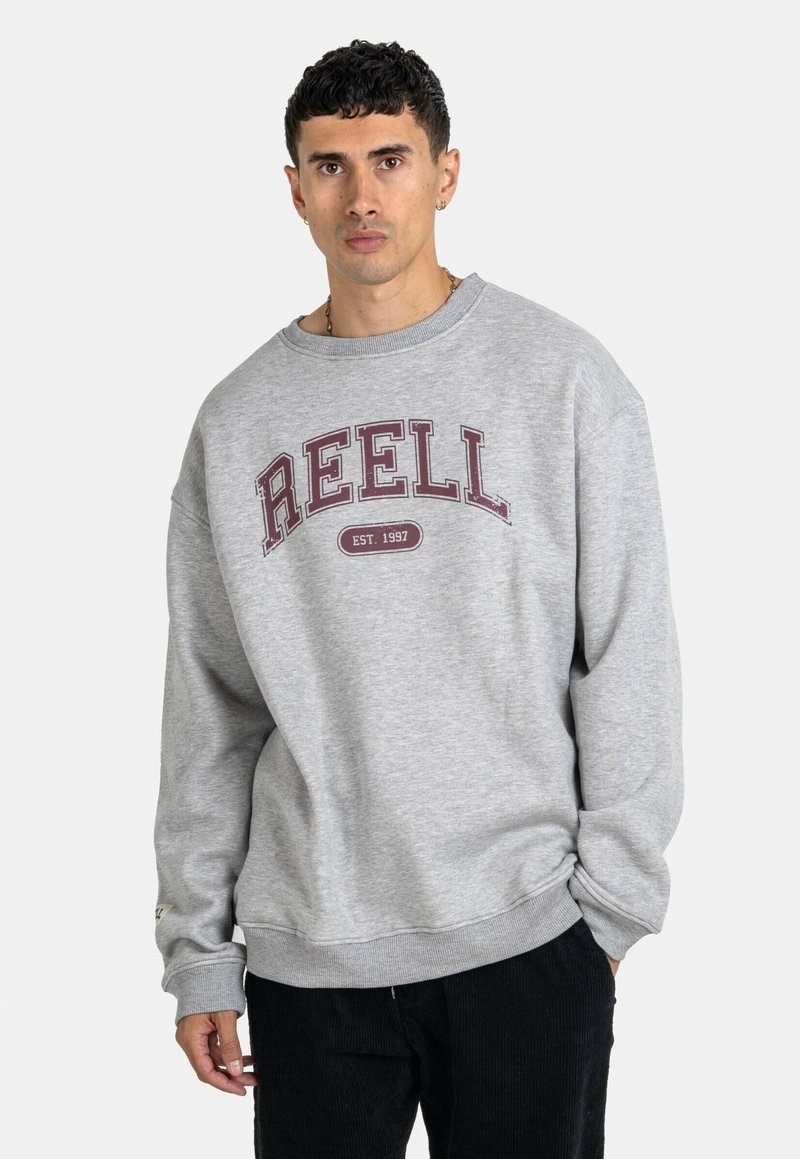 Gray sweatshirt made of soft fabric, featuring a large maroon "REELL" text and crew neckline. Accompanied by black corduroy pants.