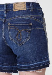 Shorts en denim bleu avec une ourlet effiloché, dotés d'un motif de couture décoratif sur la poche arrière, de quincaillerie visible et d'un patch logo en cuir.