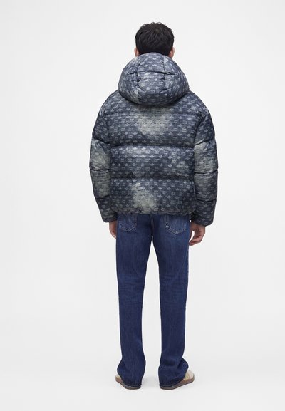Personne portant une veste matelassée bleu-gris à capuche avec un motif subtil, associée à un jean bleu foncé et des chaussures beiges, debout de dos.