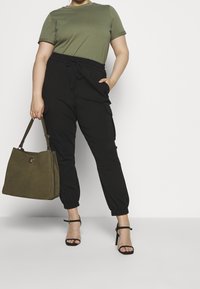 T-shirt verde a maniche corte abbinato a pantaloni cargo neri con vita elasticizzata. Il modello tiene una borsa verde oliva. Tacchi neri con cinturini completano il look.