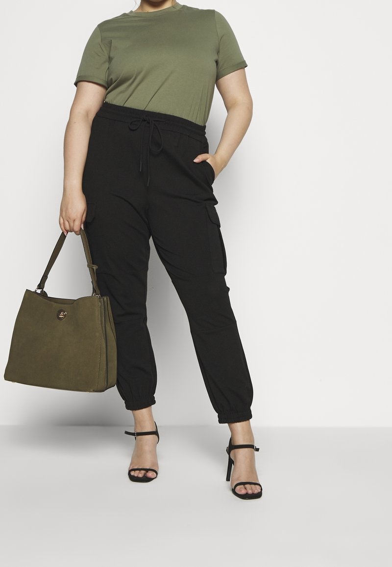 T-shirt verde a maniche corte abbinato a pantaloni cargo neri con vita elasticizzata. Il modello tiene una borsa verde oliva. Tacchi neri con cinturini completano il look.