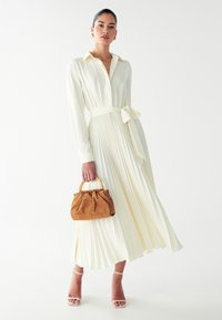 Lang geschnittenes, plissiertes Kleid in blassem Gelb mit einem Kragen und einem gebundenen Taillengürtel. Dazu eine braune Wildlederhandtasche mit einem goldenen Griff und Sandalen mit offenem Zehenbereich.