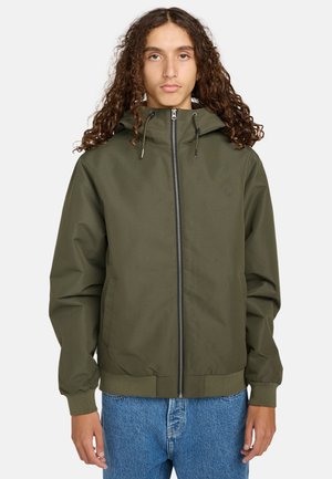 Olivengroene jas met capuchon, ritssluiting aan de voorkant, elastische manchetten en zoom, met een logo op de borst. Gemaakt over een paar blauwe jeans.