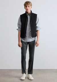 Fekete ujjatlan mellény szürke gombos ing fölött, szürke szabott nadrággal és fehér sportcipővel párosítva. Egyszerű, modern dizájn.