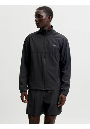 Personne aux cheveux courts portant une veste noire zippée et un short noir, regardant vers la gauche sur un fond clair uni.