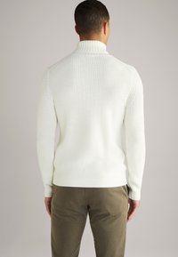 Valkoinen ribattuja neulepayejä, jossa on korkea kaulus, pitkät hihat ja tiukka muotoilu, yhdistettyinä oliivinvihreisiin housuihin, nähtynä takaapäin.