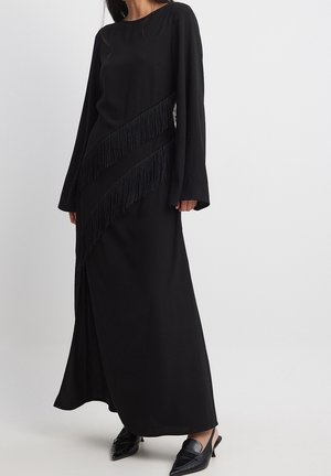 Czarna sukienka z długim rękawem, ozdobiona ukośnymi frędzlami, wykonana z miękkiego materiału, o prostej sylwetce i okrągłym dekolcie. W połączeniu z czarnymi butami.