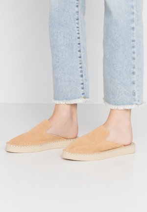 Chaussures slip-on en daim tan avec un design en V et des bords tressés, sur une semelle en caoutchouc beige clair, associées à un jean bleu clair effiloché.