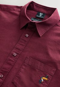 Chemise boutonnée bordeaux en coton, avec une poche poitrine ornée d'un toucan brodé et des boutons ronds.