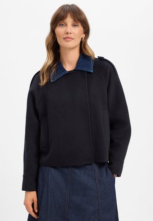 Woman wearing a black jacket with a dark blue denim collar and a matching dark blue denim skirt on a white background.