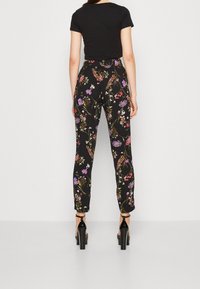 Kobieta w czarnych kwiecistych spodniach o wysokim stanie i czarnej koszulce z krótkim rękawem, stojąca tyłem do aparatu na jednolitym tle.