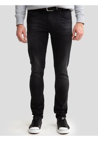 Mees, kes kannab musta vöökohaga kitsaid musti teksapükse, halli dressipluusi ja musti valgete talladega vabaajajalatsid, seisab ühevärvilise tausta ees.