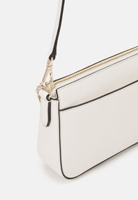 Vit läderaxelväska med svarta accenter, som har en guldfärgad dragkedja och spänne. Slät textur med minimalistisk design.