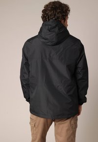 Man met krullend haar, draagt een zwart hoodie-jack en beige cargobroek, met zijn rug naar de camera gekeerd tegen een effen beige achtergrond.