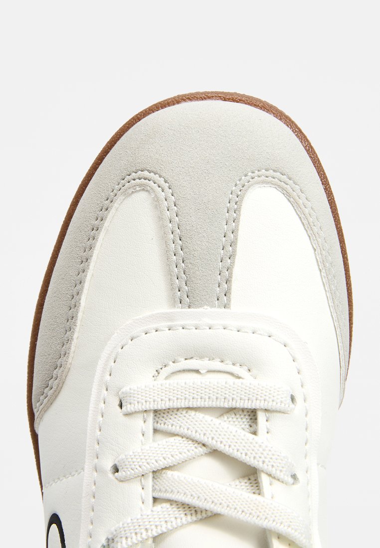 Zapatilla blanca de cuero con acentos de gamuza gris, cordones blancos lisos y suela de goma marrón. Detalle de costura a lo largo de los bordes.