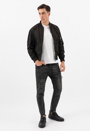 Giovane uomo in piedi con le mani nelle tasche che indossa una giacca bomber nera, una t-shirt bianca, jeans grigio scuro strappati e scarpe da ginnastica nere.