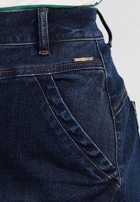 Tmavě modré džíny z denimu s vysokým pasem, viditelnými švy a značkovým kovovým štítkem na přední kapse. Hladká textura.