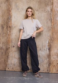 Kvinde med langt blondt hår iført en beige kortærmet skjorte, løse sorte bukser og brune sneakers, stående op ad en trævæg.