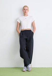 T-shirt blanche raccourcie avec le logo Nike noir, pantalon noir taille haute avec plis, et baskets Nike grises avec des accents blancs.