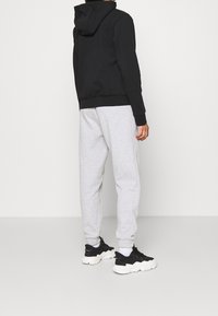 Felpa nera con tasca a marsupio frontale, pantaloni sportivi grigi e sneaker nere con suola bianca, viste da dietro su uno sfondo neutro.