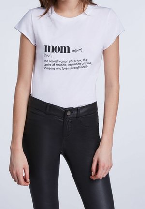 Vrouw draagt wit T-shirt met tekst die "mama" definieert als coolste vrouw, centrum van de schepping, inspiratie en onvoorwaardelijke liefde, gecombineerd met een zwarte broek.