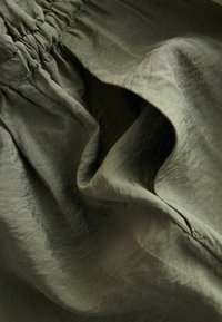 Gros plan d’un tissu vert olive avec une couture élastique froncée et des surpiqûres visibles, montrant plis et texture.