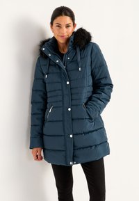 Kvinde iført en marineblå quilted vinterjakke med hætte med pelskant, lynlås og knaplukninger samt lynlåslommer i siden, stående mod en hvid væg.