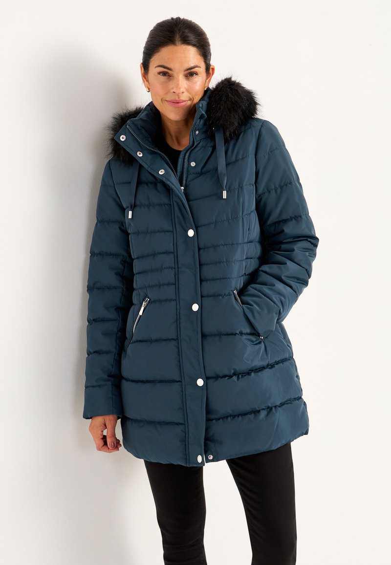 Kvinde iført en marineblå quilted vinterjakke med hætte med pelskant, lynlås og knaplukninger samt lynlåslommer i siden, stående mod en hvid væg.