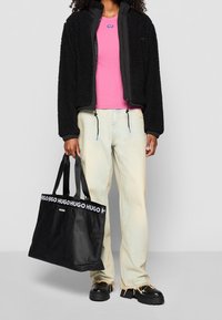 Czarna polarowa kurtka, różowa koszula, jasne dżinsy z postrzępionym dołem oraz czarna torba na ramię z białym napisem „HUGO”, noszona na czarnym obuwiu.