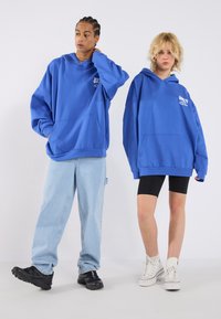 Felpe con cappuccio blu e grandi tasche, indossate da due persone. Una indossa jeans azzurri chiari, l'altra pantaloni corti neri e sneaker bianche.