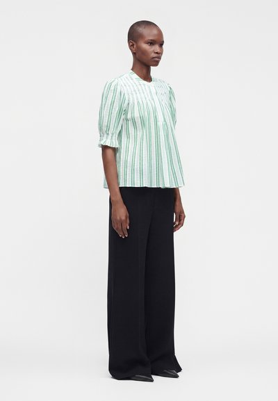 Personne debout portant un chemisier rayé vert et blanc à manches courtes et un pantalon noir à jambes larges sur un fond blanc uni.