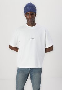 White cotton t-shirt featuring a small black script design reading “Edition,” paired with light blue denim jeans and a blue ribbed beanie.