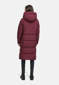 Cappotto lungo burgundy imbottito con cappuccio, caratterizzato da una texture trapuntata, tasche laterali e vestibilità comoda, indossato sopra leggings neri e stivali.
