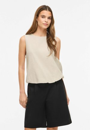 Jeune femme aux longs cheveux bruns portant un haut beige sans manches et un short noir ample longueur genoux sur fond blanc uni.