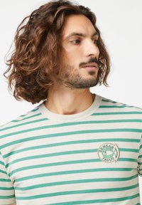 Baumwoll-T-Shirt mit cremefarbenem Hintergrund und grünen horizontalen Streifen. Verfügt über einen Rundhalsausschnitt und eine kreisförmige Logo-Stickerei auf der Brust.