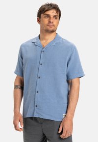 Krátky rukáv, svetlomodrá košeľa na gombíky vyrobená z textúrovanej tkaniny, s rozšíreným golierom a hnedými gombíkmi pozdĺž prednej strany.