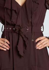 Burgunder klänning med volangkrage och knytmidja. Har ett justerbart bälte med silveröglor, dekorativa kristallaccent och strukturerad tyg.