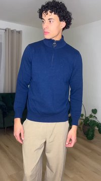 Nooruk lokkis juustega mees, kes kannab tumesinist lukuga kampsunit ja beeže pükse, seisab siseruumides roheliste taimede ja diivani lähedal.