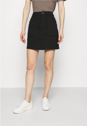 Femme portant une mini-jupe en jean noire avec des poches avant, un haut marron et des baskets blanches, debout sur un fond clair uni.