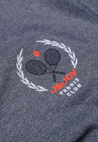 Donkergrijze stof met een gestructureerde afwerking toont een witte lauwerkrans en zwarte tennisrackets die zijn geborduurd, verfraaid met een rode tennisbal en tekst.