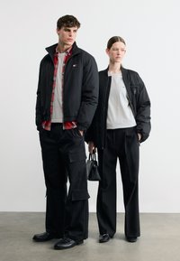Νεαρός άνδρας και γυναίκα στέκονται, και οι δύο φορούν μαύρα μπουφάν, μαύρο παντελόνι και γκρι μπλουζάκια· η γυναίκα κρατά μια μικρή μαύρη τσάντα στο πλάι.
