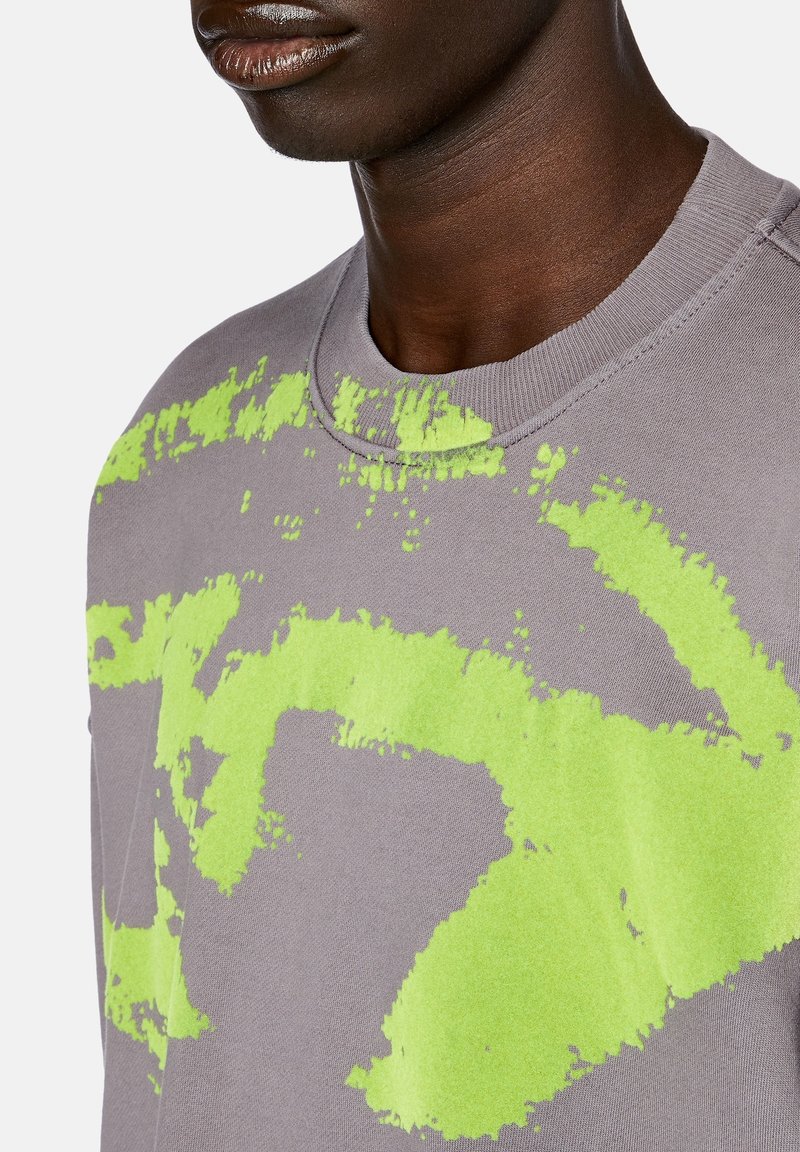 Sweatshirt gris avec un motif éclaboussé graphique en vert vif. Encolure ronde et coutures visibles. Tissu doux et texturé.