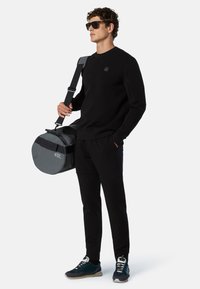 Felpa nera e pantaloni indossati con occhiali da sole. Tiene una borsa da viaggio grigia e rotonda con la scritta bianca "45L". Le scarpe da ginnastica presentano dettagli blu navy e neri.