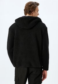 Chaqueta de forro polar negra, texturizada, con capucha, que presenta un corte holgado y mangas largas. La tela tiene una apariencia suave y esponjosa.