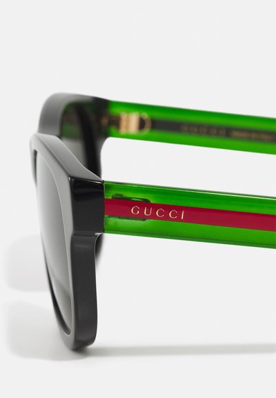 Juodos stačiakampės saulės akinių rėmeliai su žaliai skaidriomis rankenomis, kuriose yra raudona juostelė ir aukso spalvos "GUCCI" logotipas šone.
